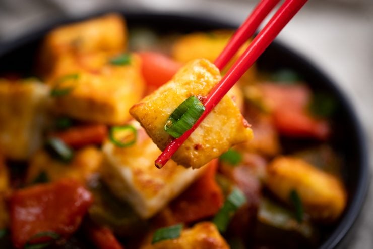 Chop sticks holding up a piece of paneer in front of a bowl of chili paneer