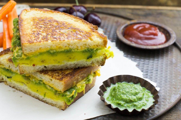 Masala cheese toast sandwich halves on a round tray atop white paper, served with carrot sticks, cherries, chutney and ketchup