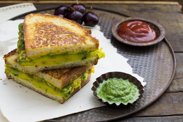 masala cheese toast sandwich halves layered one above another with cherries, ketchup and chutney on white paper and round tray